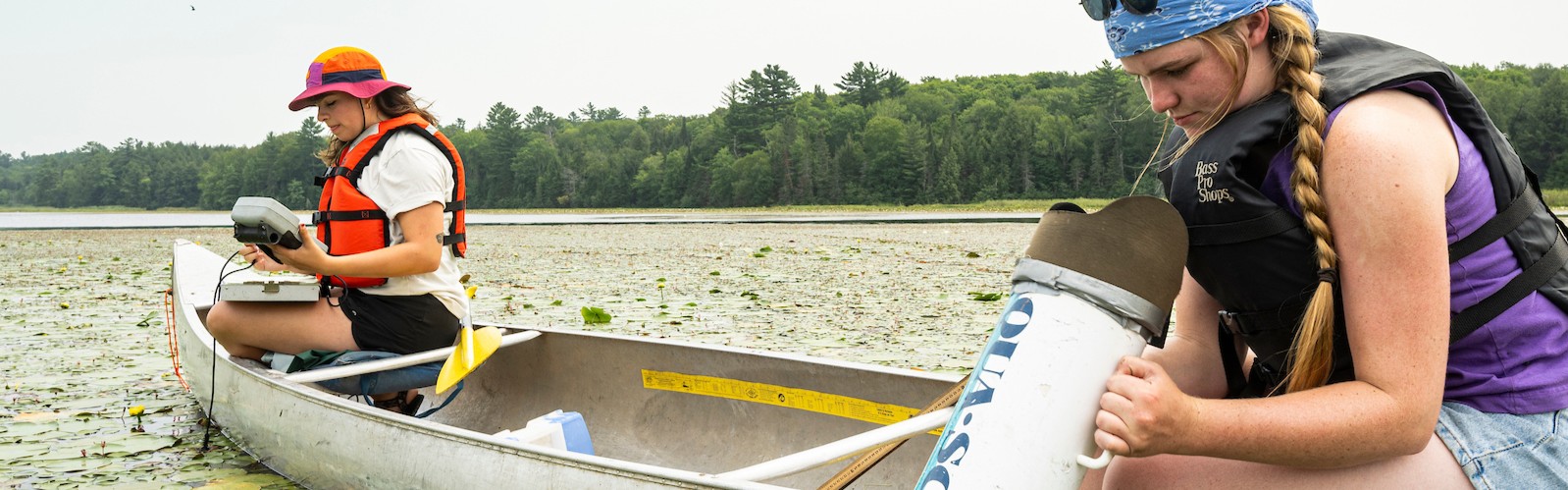 Two people sitting in a canoe on a lake covered by lily pads. One dressed in an orange hat a life jacket, the other in a blue bandana and black vest -both are taking measurements
