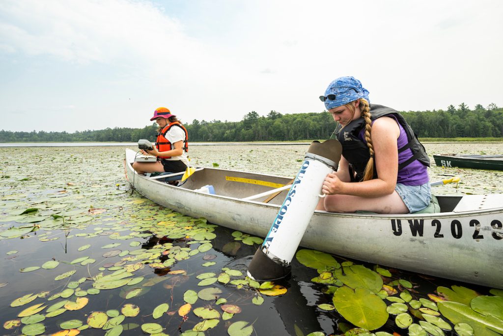 Two undergraduate researchers in a canoe on a lake surrounded by lily pads, taking measurements. One student wearing an orange lifejacket and sun hat, the other in a black lifejacket and blue bandana.
